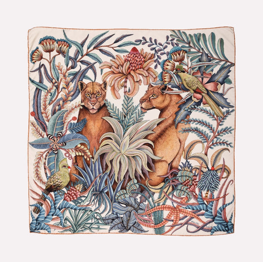 A silk scarf featuring an intricate African wildlife scene with two lionesses resting among lush flora, a vibrant bird, and winding vines in soft earth tones.