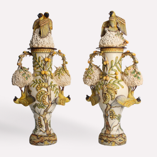 A pair of grand urns adorned with intricate sculpted masked weaver birds and woven nests. The urns are embellished with ochre and yellow tones, symbolising abundance and nourishment.