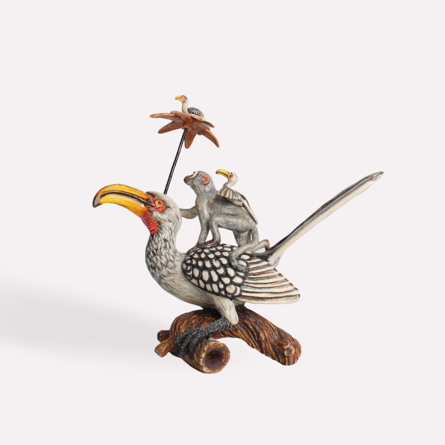 A ceramic sculpture of a black-and-white hornbill with a curved yellow beak, perched on a wooden-like base. A small gray monkey sits on the bird's back, holding a miniature umbrella with a tiny bird perched on top.