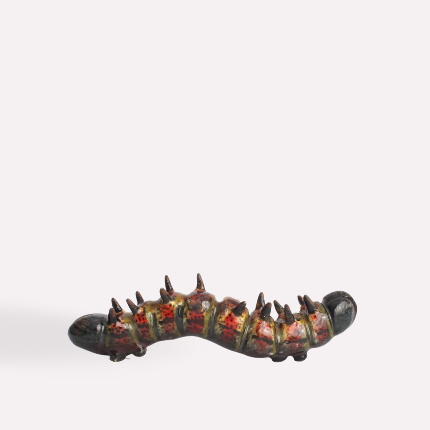 A hand-painted ceramic caterpillar sculpture with a curved body, dark green base, and intricate yellow and red detailing, featuring small black spines along its back.