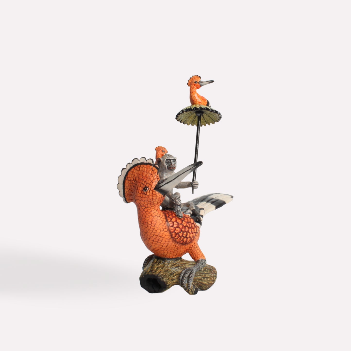 A ceramic sculpture featuring a vibrant orange hoopoe bird with black and white wings, standing on a textured base. A small monkey sits atop the bird, holding a tiny green umbrella with another miniature hoopoe perched on top.