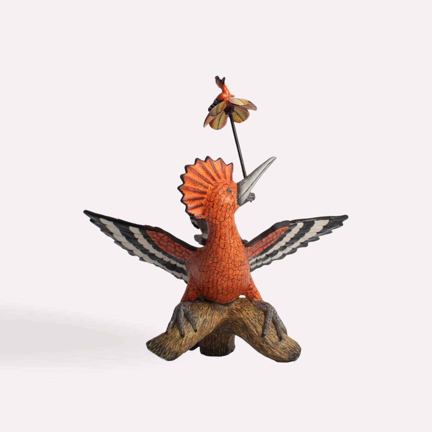 A detailed ceramic sculpture of a vibrant orange hoopoe bird with its wings spread wide, perched on a naturalistic base, carrying a small monkey figure on a thin pole above its head.