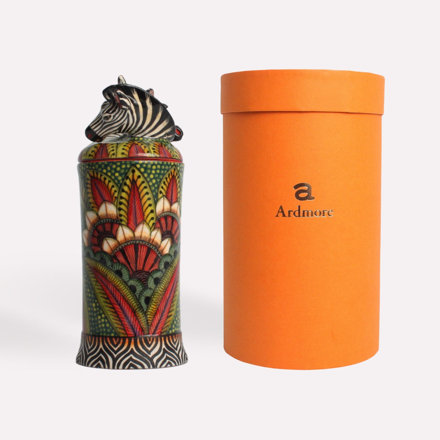 A tall, hand-painted ceramic diffuser pot featuring intricate African-inspired patterns in red, green, and gold, alongside a sleek orange container for packaging.