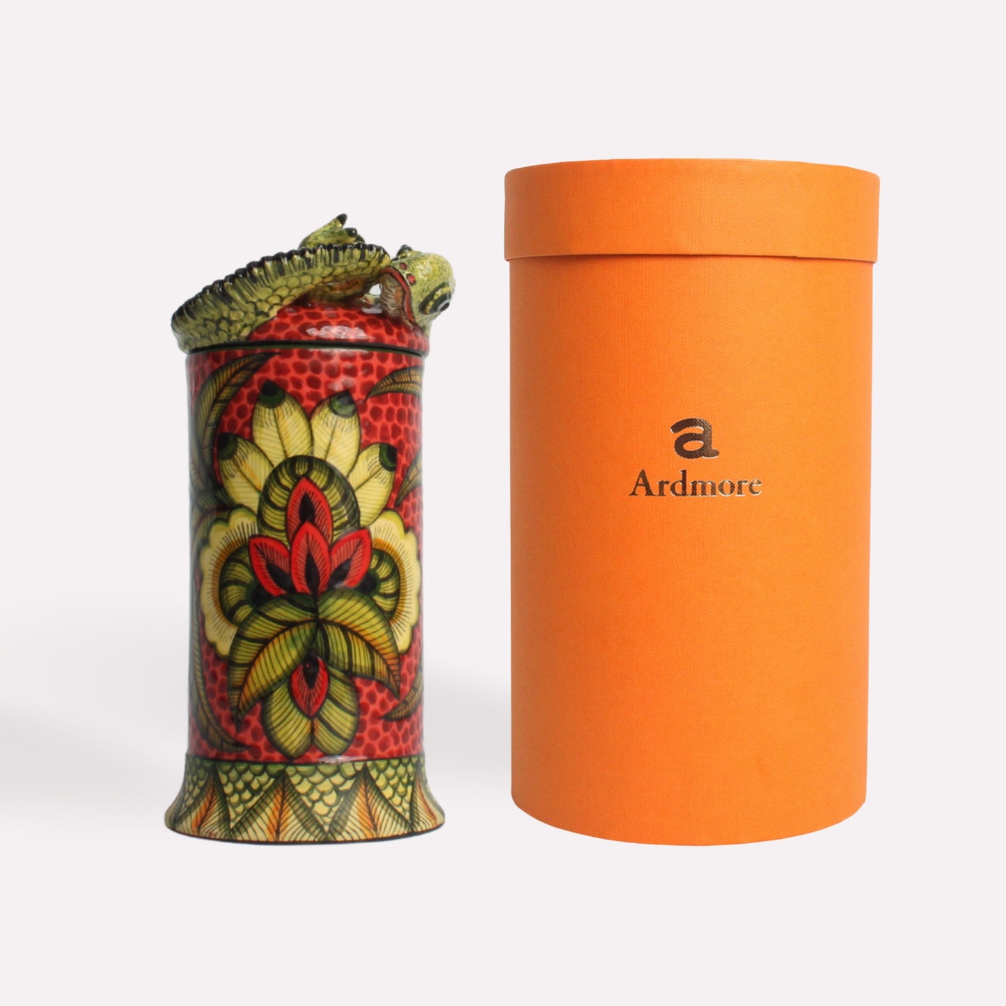 A cylindrical ceramic diffuser pot with intricate red, green, and yellow chameleon-themed designs, placed beside an orange Ardmore-branded packaging tube.