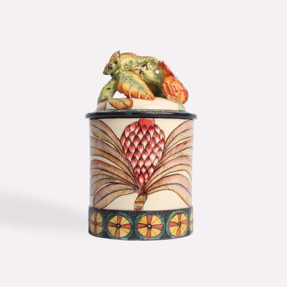 A close-up of the ceramic candle jar, highlighting the detailed chameleon figurine resting on the lid. The body of the jar is adorned with elegant geometric patterns and earthy colours.