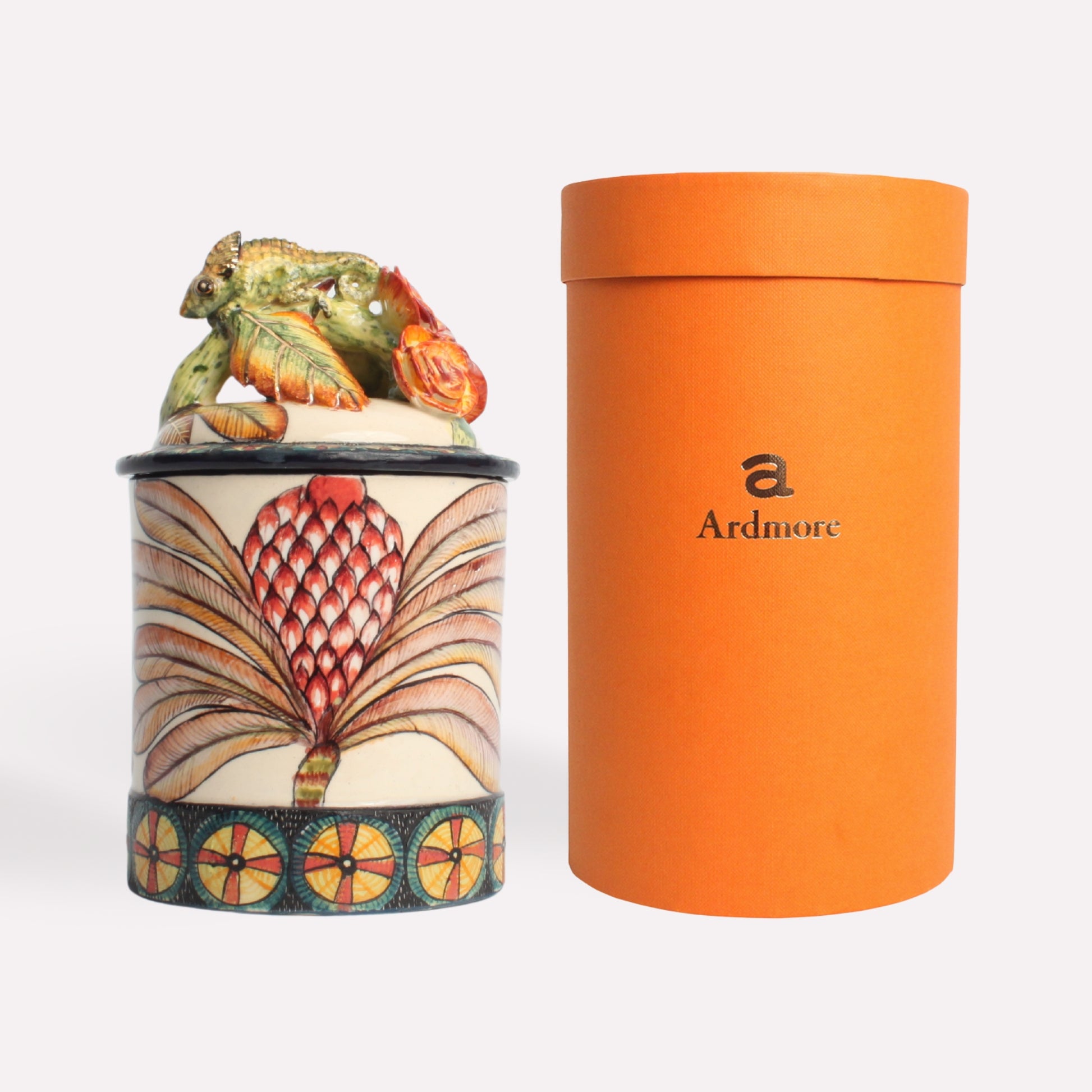 A hand-painted ceramic candle jar with a sculpted chameleon perched on the lid, accompanied by a tall, sleek orange container for packaging. The jar features intricate African-inspired patterns in red, gold, and cream tones.