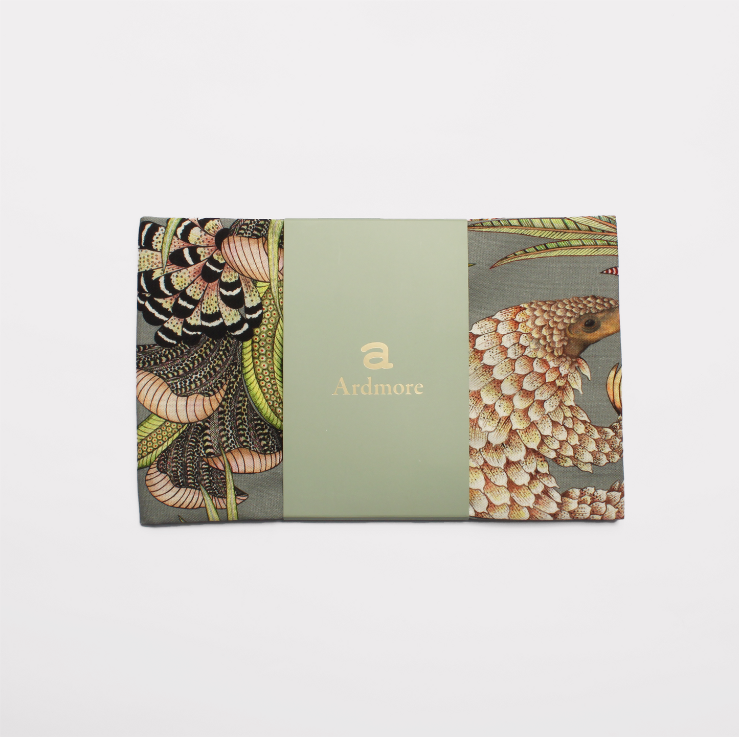 A packaged tea towel with a visible preview of the pangolin and botanical artwork, set against a dark fabric background.