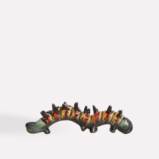 A hand-painted ceramic caterpillar sculpture with a curved body, dark green base, and intricate yellow and red detailing, featuring small black spines along its back.