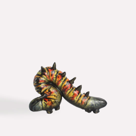 A hand-painted ceramic caterpillar sculpture with a curved body, dark green base, and intricate yellow and red detailing, featuring small black spines along its back.