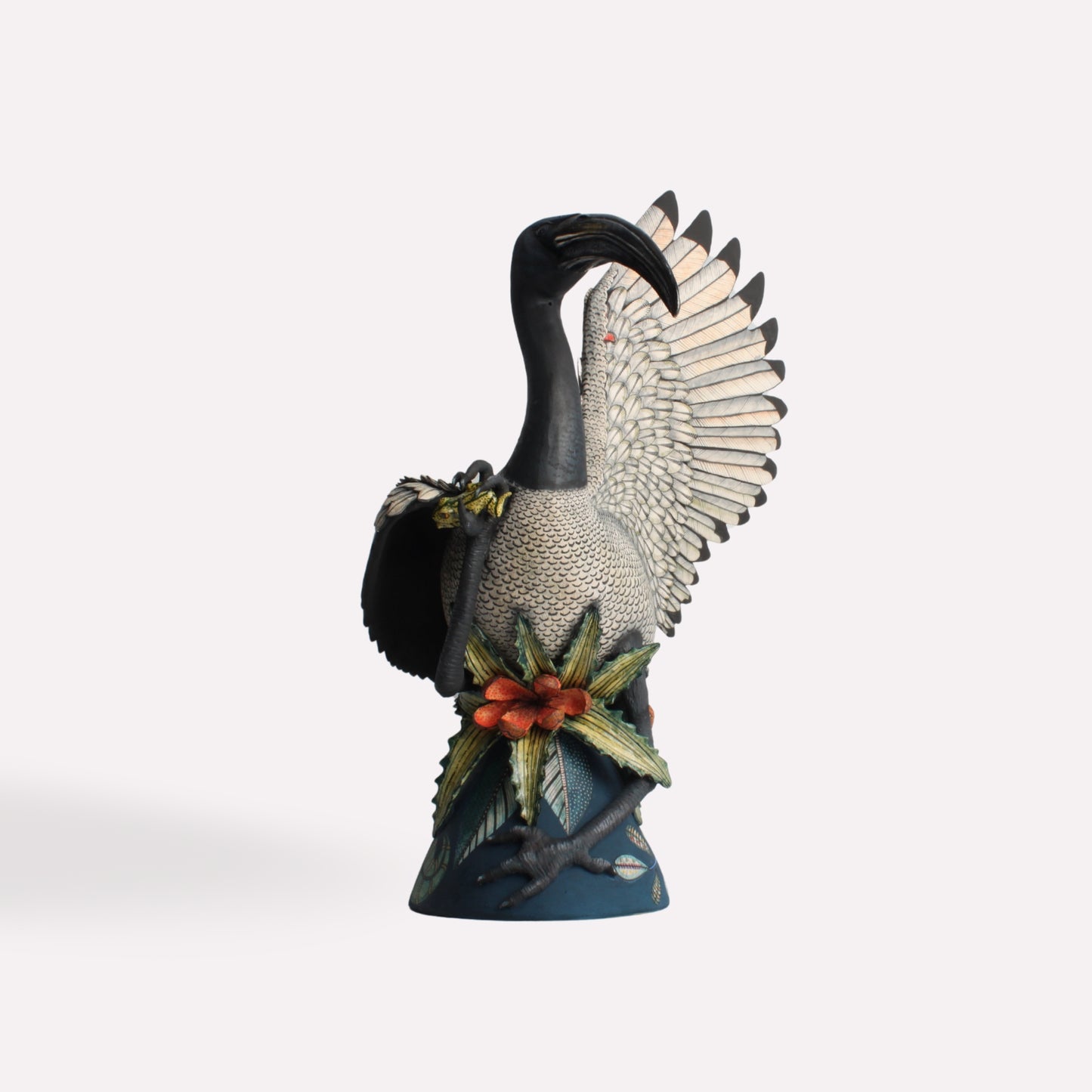 A meticulously crafted ceramic sculpture of an African Sacred Ibis standing gracefully with its wings partially extended. The bird’s black and white plumage is intricately detailed, with subtle shading and texture enhancing its realism. The base is adorned with floral elements, adding a vibrant contrast.