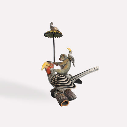 A hand-painted ceramic sculpture featuring a black-and-white hornbill perched on a log, with a small monkey figure sitting on its back, holding onto a tall pole with a decorative parasol and a tiny bird on top.