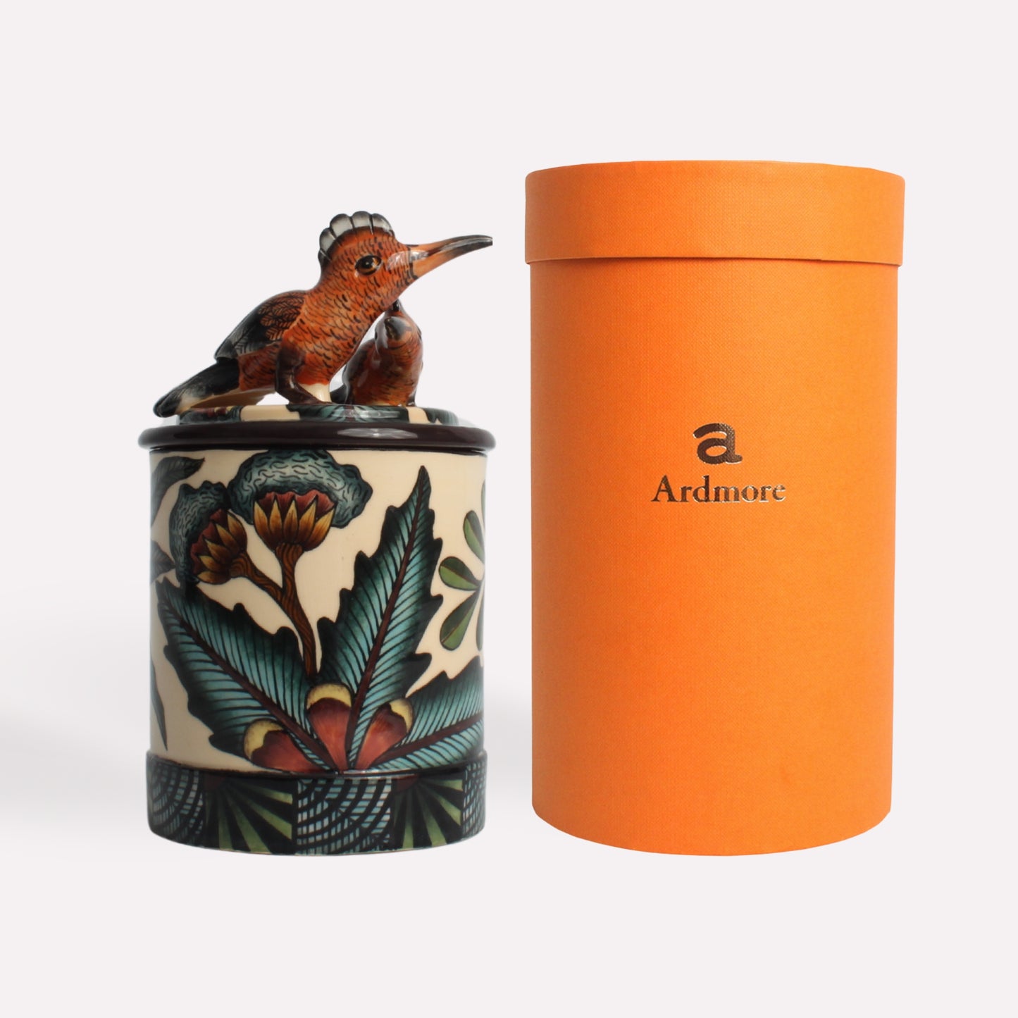 A handcrafted ceramic candle container featuring a sculpted hoopoe bird perched on the lid, paired with an orange Ardmore-branded packaging tube.