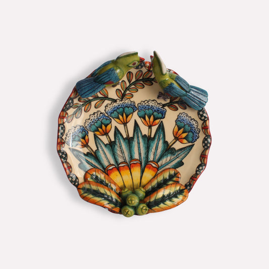 A round ceramic coin dish featuring a hand-painted turaco bird at the centre, surrounded by lush green foliage and an orange floral pattern.