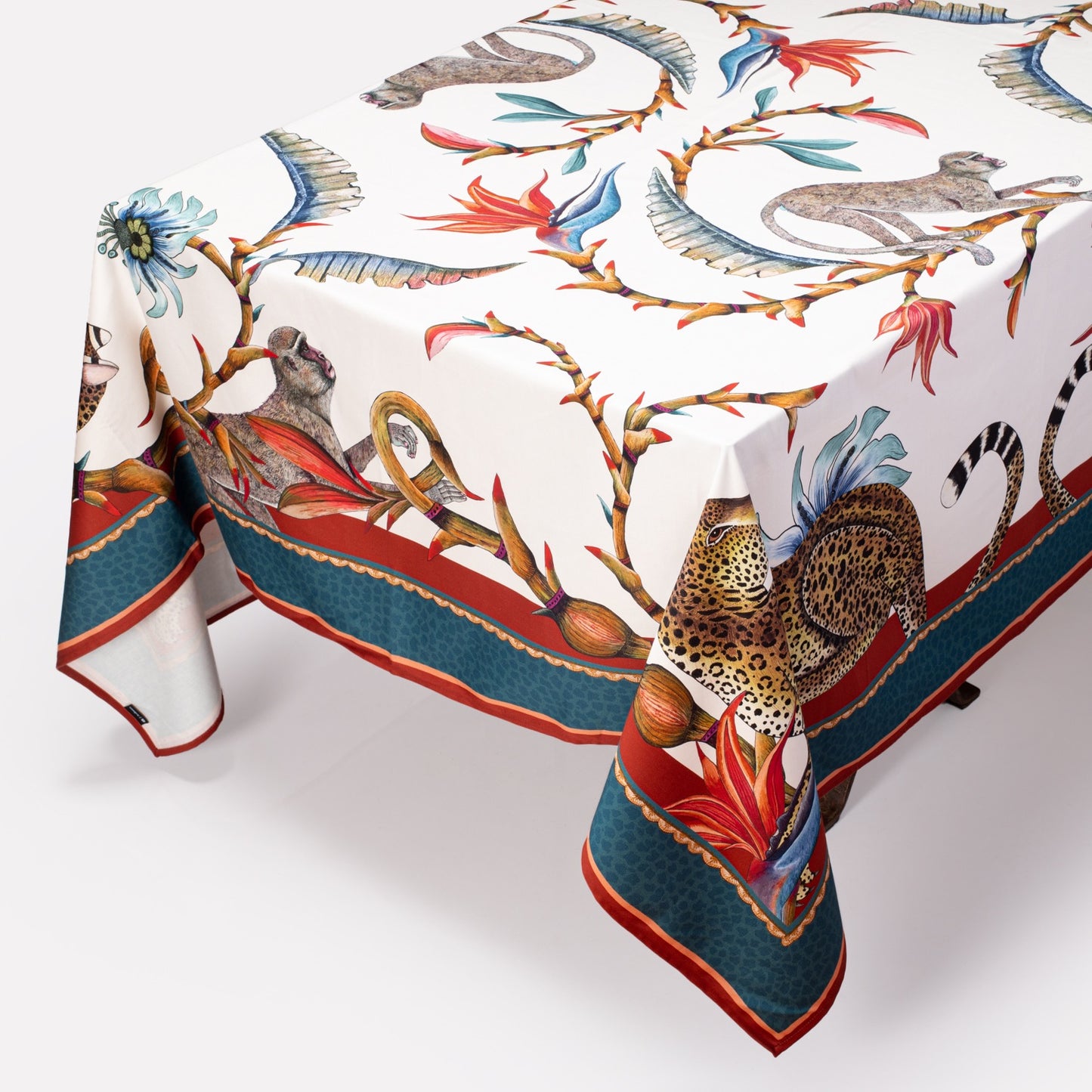 A square tablecloth with a white base, featuring an intricate design of monkeys, leopards, birds of paradise, and passion flowers, bordered in red and blue.